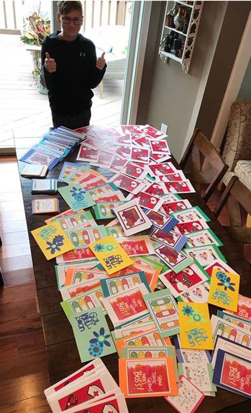 One boy stands with two thumbs up in front of hundred of homemade cards on a table. 